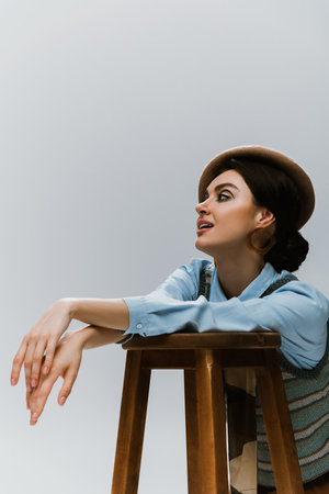 young stylish woman in beret and autumnal clothes leaning on wooden high chair isolated on greyの写真素材