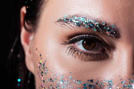 close up view of young woman with sparking glitter on eyebrow and face isolated on black