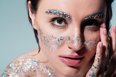 portrait of brunette woman with glitter on cheeks and eyebrows looking at camera isolated on grey