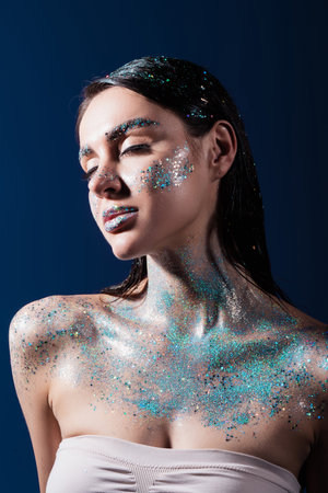 portrait of brunette young woman with closed eyes and glitter on body isolated on blue