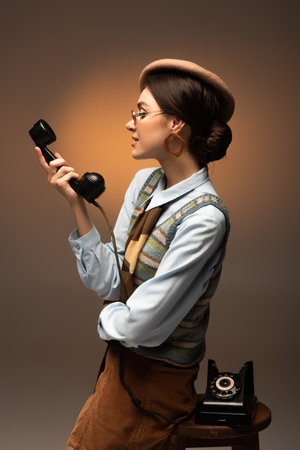 stylish young woman in beret and sunglasses looking at vintage telephone on brown