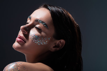 portrait of young woman with closed eyes and glitter on eyebrows isolated on grey