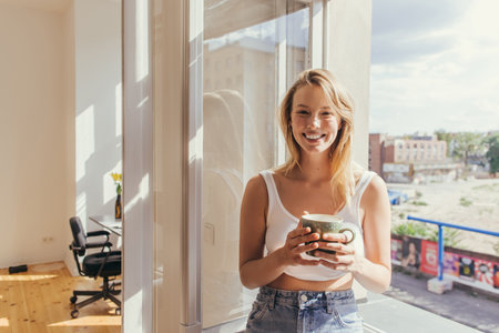 Happy blonde woman in top holding cup and looking at camera near open window at home