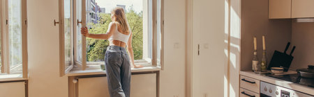 Young woman looking at open window near cup on windowsill in kitchen, bannerの写真素材