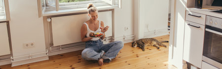 Cheerful woman holding plate with delicious breakfast near scottish fold cat on floor at home, bannerの写真素材