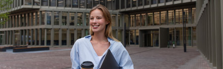Young freelancer in shirt holding takeaway drink and laptop outdoors in Berlin, banner