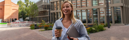 Cheerful blonde freelancer holding paper cup and laptop on urban street in Berlin, banner