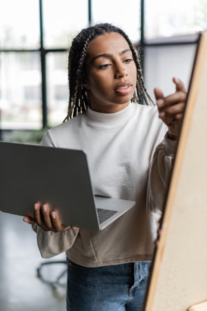 African american businesswoman holding laptop near blurred board in office