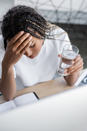 High angle view of tired african american businesswoman holding glass of water near computer in...