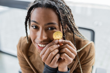 KYIV, UKRAINE - APRIL 27, 2022: Smiling african american businesswoman holding bitcoin and looking...