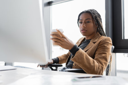 African american businesswoman holding coffee to go near computer in officeの写真素材