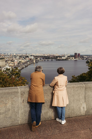 back view of retired couple in beige coats standing on bridge near river with city view