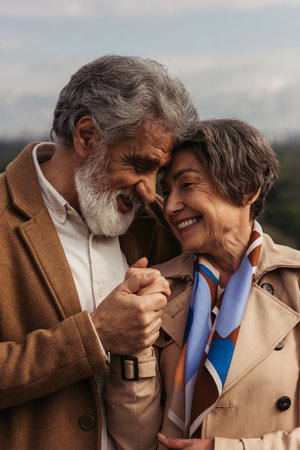 cheerful man smiling while hugging and holding hands with senior wife in trench coat