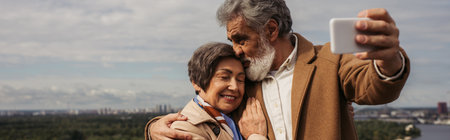 bearded senior man kissing head of cheerful wife and taking selfie, banner