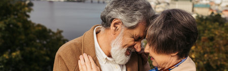 senior woman hugging with bearded husband with grey hair and smiling near river, banner