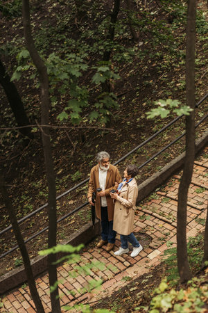 high angle view of joyful retired couple in beige coats holding paper cups and walking in parkの写真素材