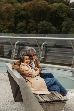 happy senior man hugging elderly wife while sitting on bench near bridge guard railの写真素材