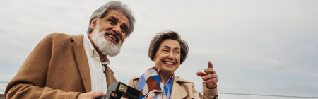 bearded and senior man holding vintage camera near wife smiling while pointing with finger, bannerの写真素材