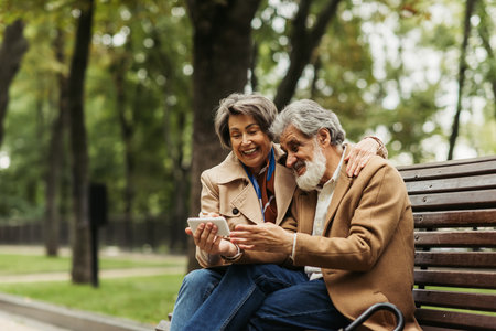 happy senior couple in coats sitting on bench and looking at smartphone