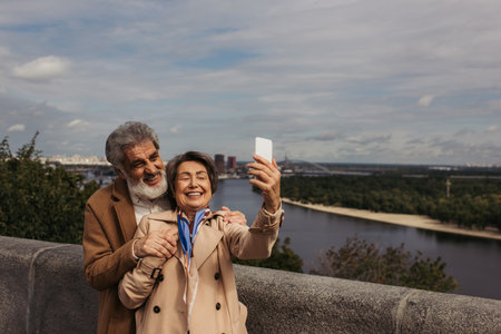 happy senior woman in beige trench coat and taking selfie with bearded husband