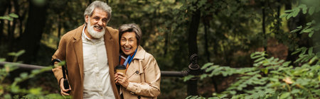 pleased senior couple in beige coats holding coffee to go in autumnal park, banner