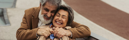 cheerful senior man in coat hugging elderly wife smiling with closed eyes