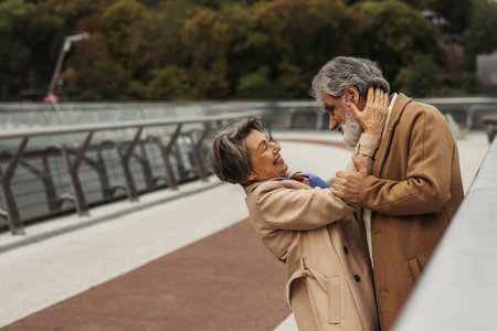cheerful elderly woman hugging bearded husband in beige coat on bridge near guard rail