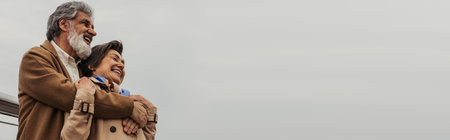 low angle view of happy senior man hugging positive wife in beige coat and smiling outside, banner