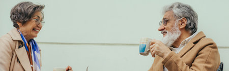 cheerful senior woman in eyeglasses and trench coat smiling near happy husband holding cup with tea, bannerの写真素材