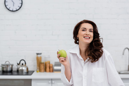 Smiling woman in shirt holding fresh apple in kitchen