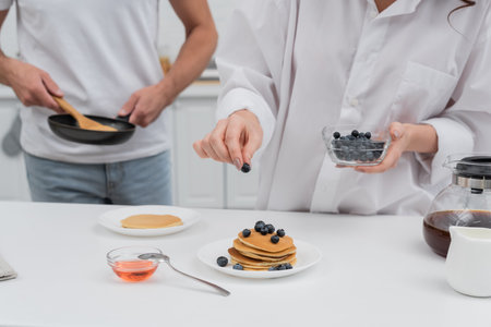 Cropped view of woman holding blueberries near pancakes and blurred husband with frying pan in kitchenの写真素材