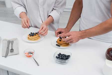 Cropped view of couple putting fresh blueberries on pancakes near honey on table in kitchen