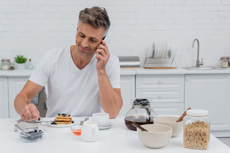 Smiling man talking on smartphone near delicious breakfast in kitchenの写真素材