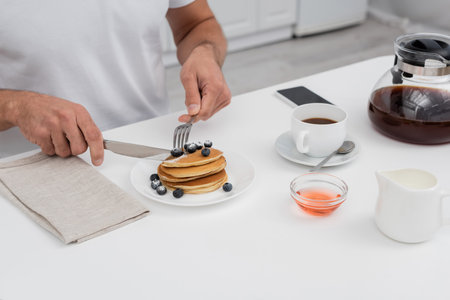 Cropped view of man cutting pancakes near coffee and smartphone in kitchen