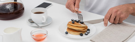 Cropped view of man cutting pancakes with blueberries near coffee and smartphone, bannerの写真素材