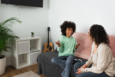 curly african american girl smiling while gesturing near mother in living roomの写真素材