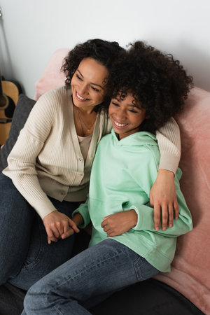 high angle view of happy african american mother hugging cheerful preteen daughter in living room