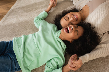top view of happy african american girl laughing while lying on carpet with cheerful motherの写真素材