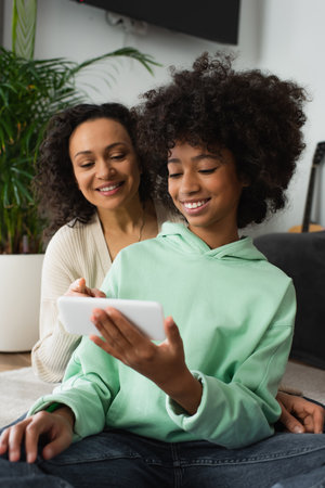 happy african american preteen girl using mobile phone near positive motherの写真素材