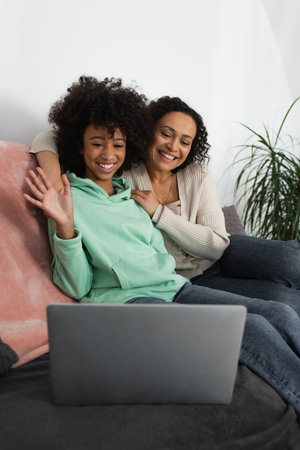 happy african american mother hugging cheerful preteen daughter waving hand during video call