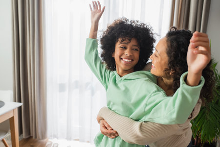 cheerful african american woman hugging happy preteen daughter with raised handsの写真素材