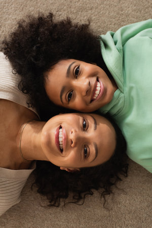 top view of happy african american girl lying on carpet with cheerful mother and looking at camera