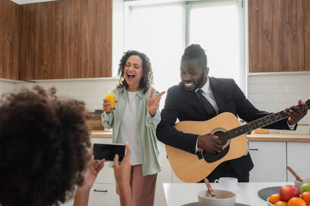 curly african american kid taking photo of happy father in suit playing acoustic guitar near mother...