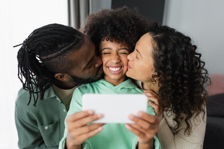 happy african american parents kissing cheek of daughter while taking selfie at homeの写真素材