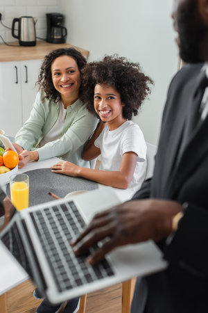 cheerful african american mother holding smartphone near daughter and looking at husband in suit using laptop on blurred foregroundの写真素材