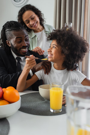 happy african american parents smiling and looking at preteen daughter eating corn flakes in kitchen