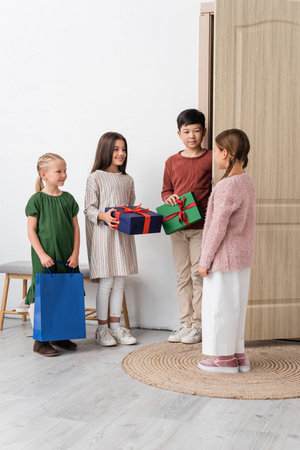 Cheerful interracial kids holding presents near friend and door at homeの写真素材