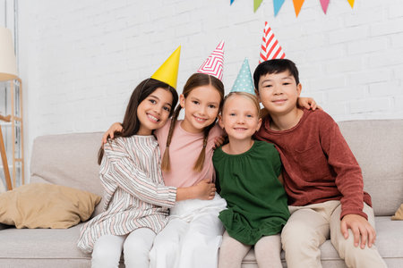 Smiling interracial kids in party caps looking at camera and hugging during birthday party at homeの写真素材