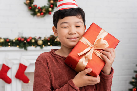 Happy asian boy in party cap holding gift near blurred christmas decor at homeの写真素材