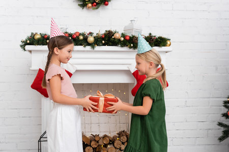 Side view of girl in party cap giving present to friend near christmas decor on fireplace at homeの写真素材